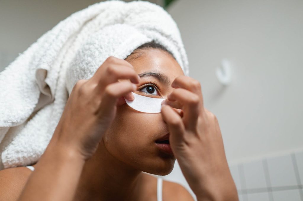 A woman with a towel on her head applies under eye masks during her skincare routine.